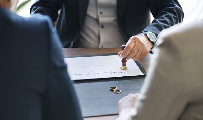 Une main apposant un cachet sur un certificat de divorce, avec des alliances posées sur le bureau, en présence de deux personnes formellement habillées.