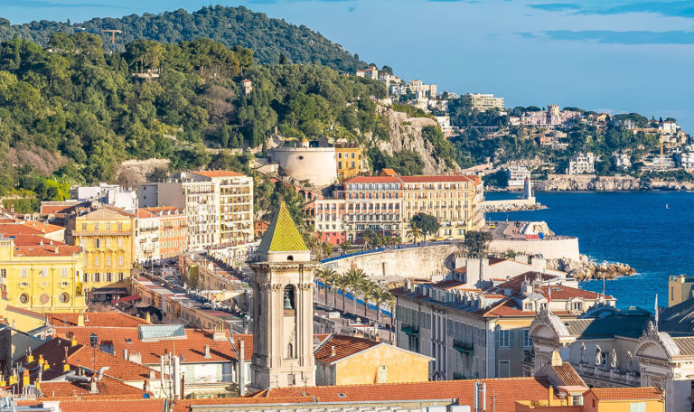Vue panoramique des toits de la ville de Nice, avec des bâtiments colorés, des collines verdoyantes et la mer Méditerranée en arrière-plan sous un ciel bleu éclatant.