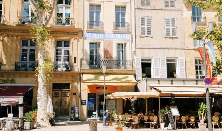 Façades ensoleillées de bâtiments à Aix-en-Provence, avec des terrasses de cafés animées et des devantures colorées, un cadre discret pour des observations en milieu urbain.