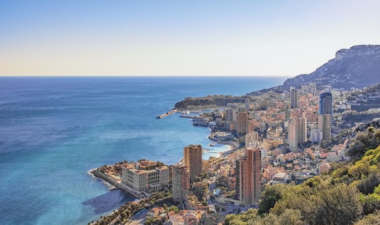 Vue panoramique sur Monaco et ses environs, avec ses bâtiments modernes en bord de mer et les montagnes en arrière-plan, idéal pour des investigations discrètes dans un cadre luxueux.