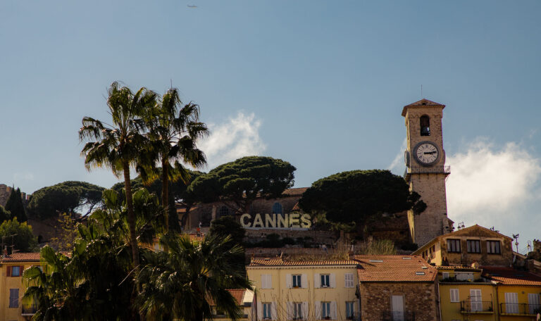 Vue sur la célèbre inscription "Cannes" et le clocher d'une église sur une colline, avec des toits de maisons provençales et des palmiers en premier plan, idéal pour des enquêtes discrètes dans un cadre emblématique.